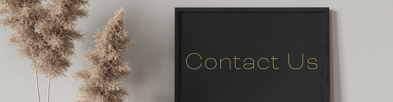 A framed 'Contact Us' sign in gold lettering on a black background, hung on a wall beside pampas grass.